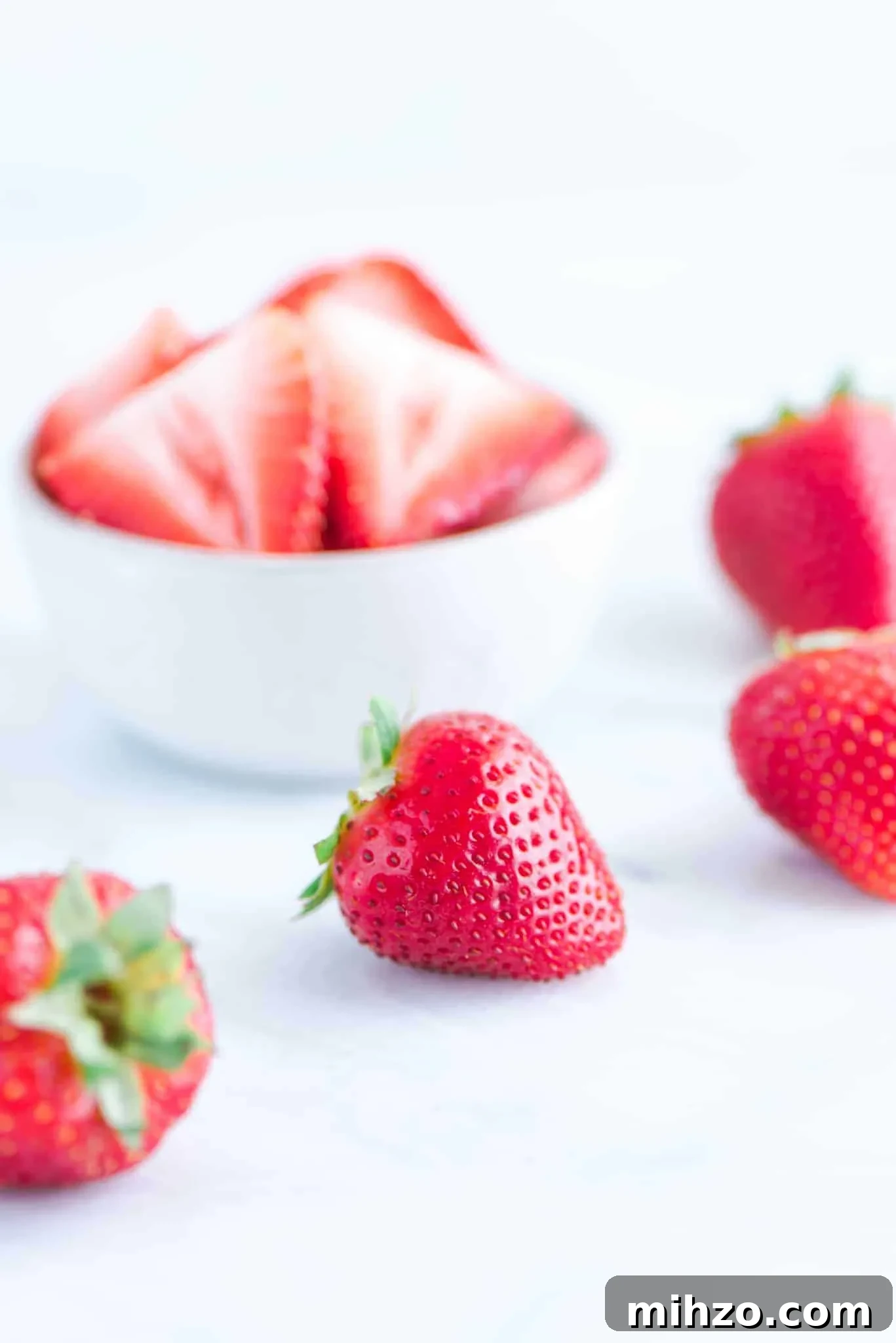 Gluten-Free Strawberry Icebox Cake Recipe for Summer Parties 3 sliced strawberries in a white bowl with whole strawberries on a marble slab
