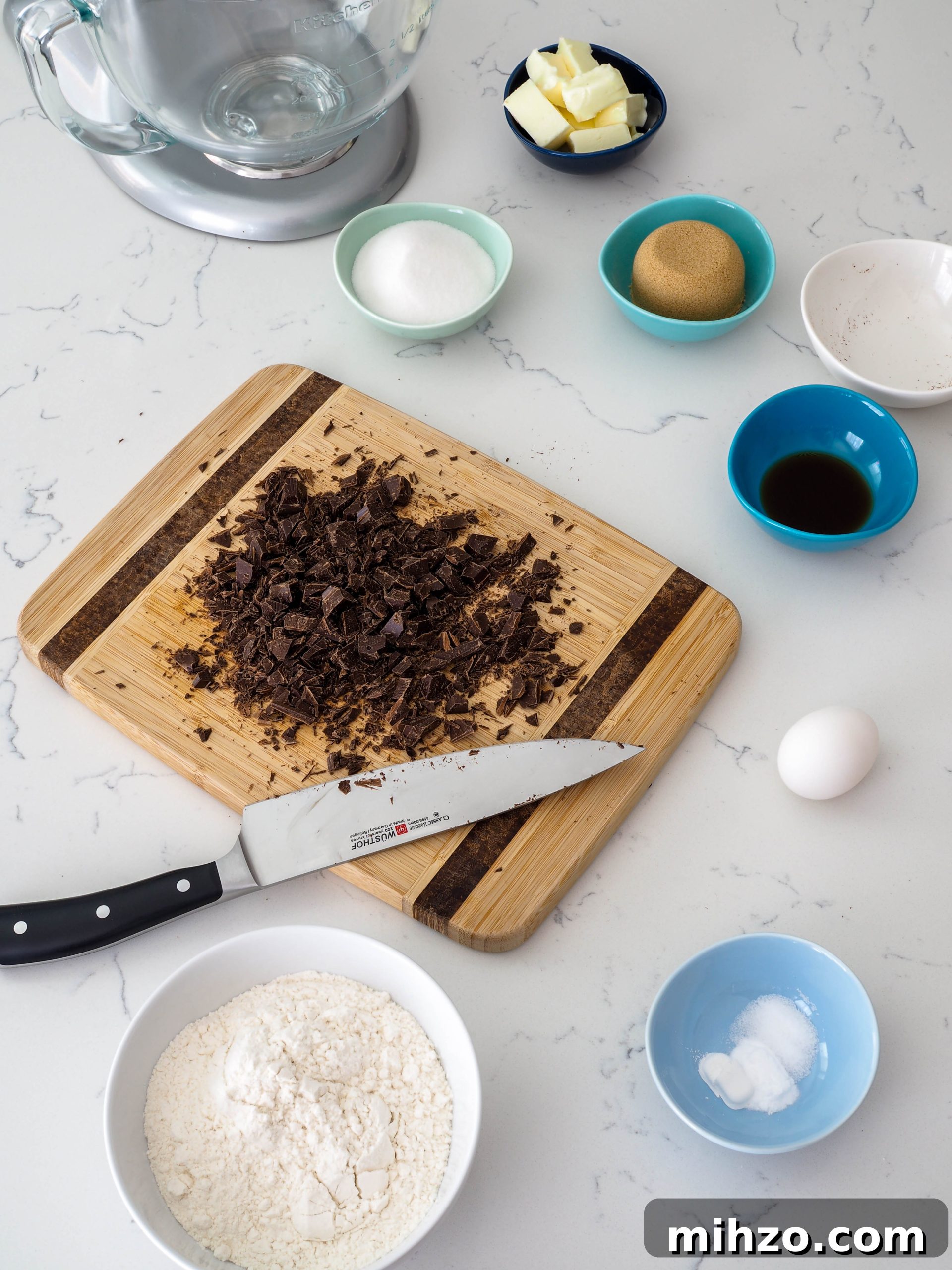 A cutting board covered with roughly chopped chocolate and a Wusthof chef's knife is surrounded by cookie ingredients.