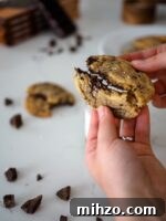 A dark chocolate chip cookie is broken in half by two hands.