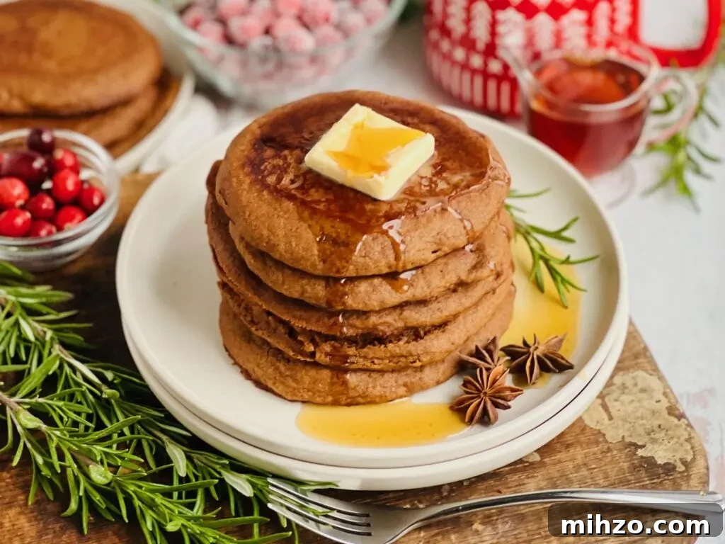 Gingerbread Pancakes Recipe: Fluffy Spiced Holiday Pancakes 4 A white plate filled with dark brown colored round pancakes with a large square of melty butter and syrup running down the top and a small bowl of cranberries and syrup nearby.