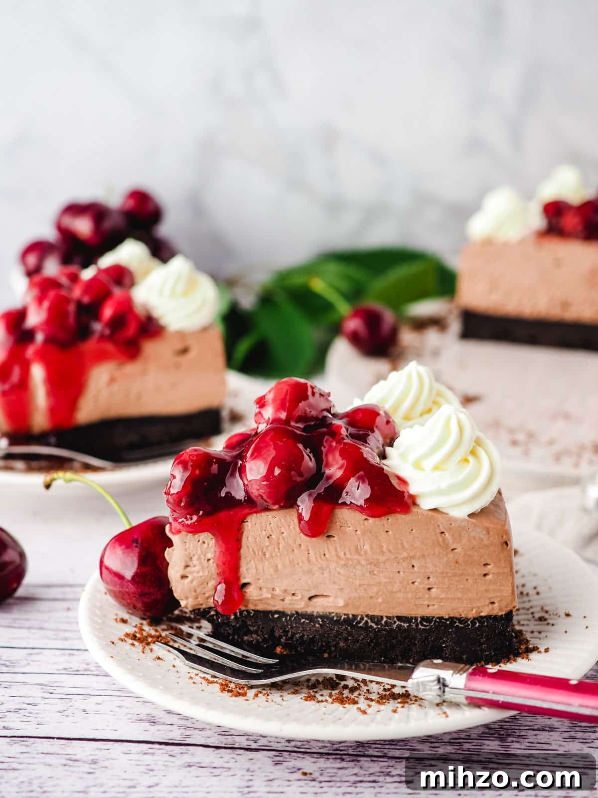 Slice of black forest gateau cheesecake on a plate with a fork and rest of cheesecake in the background.