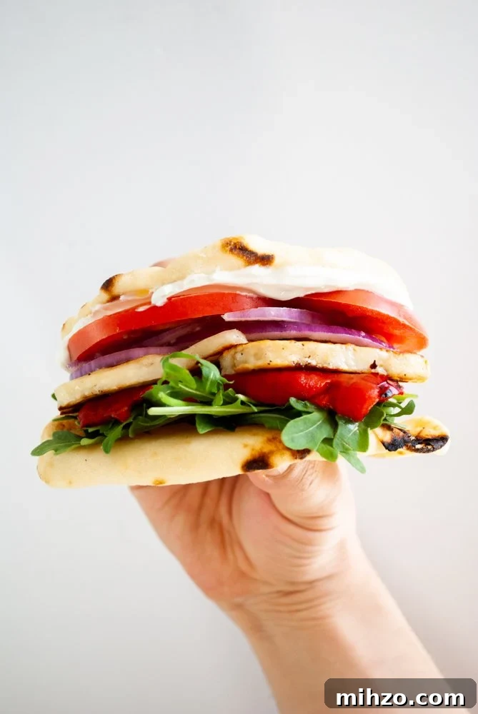 A hand holding a sandwich of fried halloumi with roasted red pepper, arugula, tomato, red onion and tzatziki.