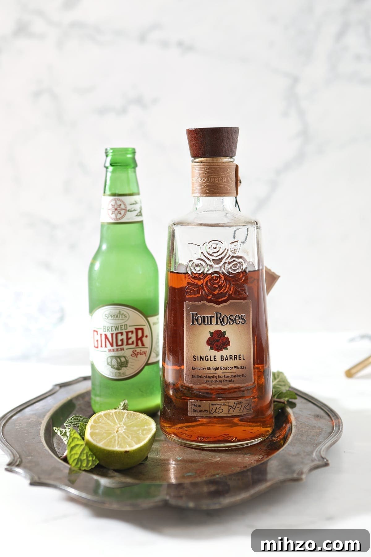 A bottle of bourbon, ginger beer and a lime with mint are shown on a silver platter