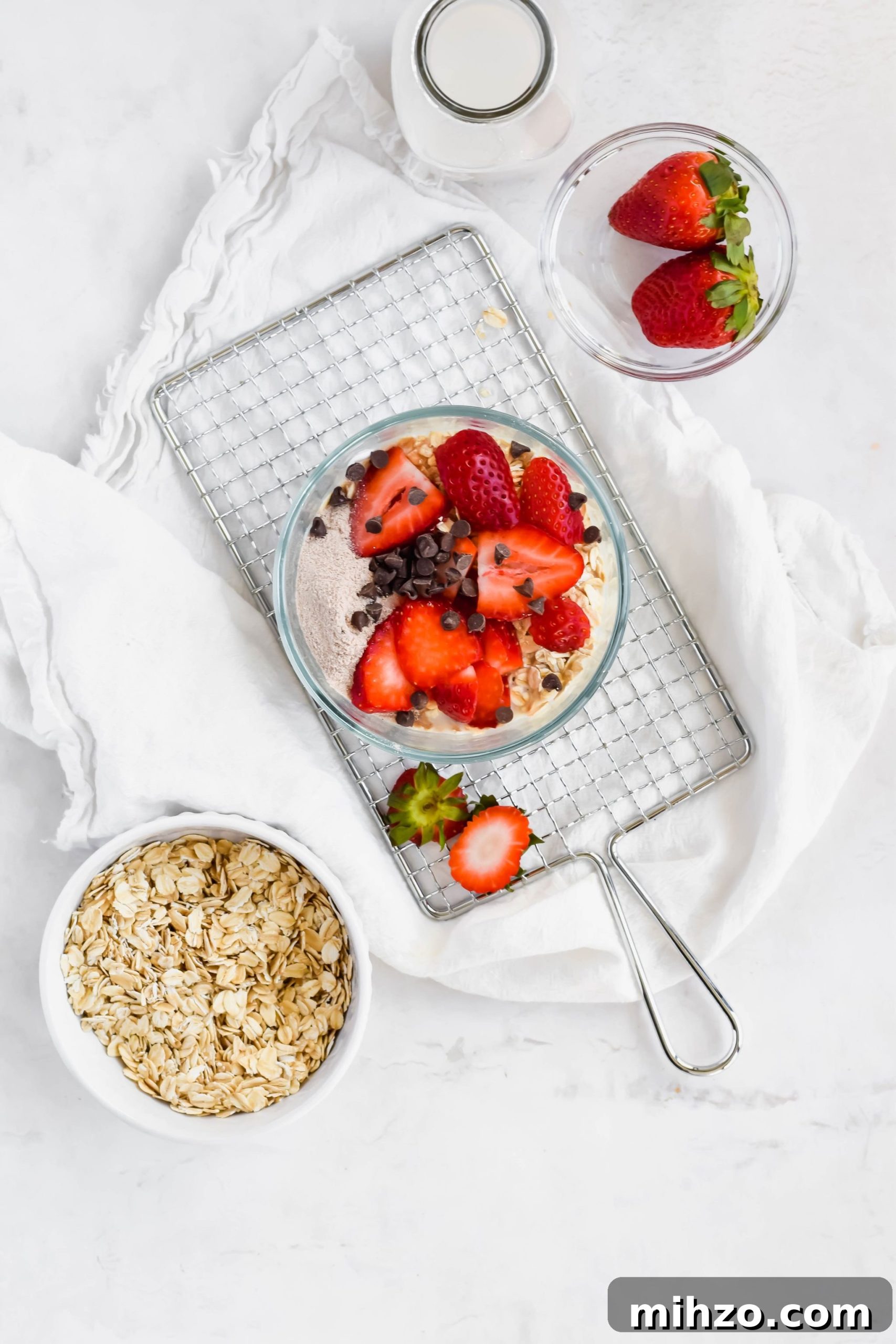 Chocolate-Strawberry Overnight Oats Recipe for Creamy Mornings 6 Strawberry Chocolate Overnight Oats ingredients in a small glass bowl next to a bowl of rolled oats, a bowl of fresh strawberries, a bowl of mini chocolate chips, and a tall milk jug.