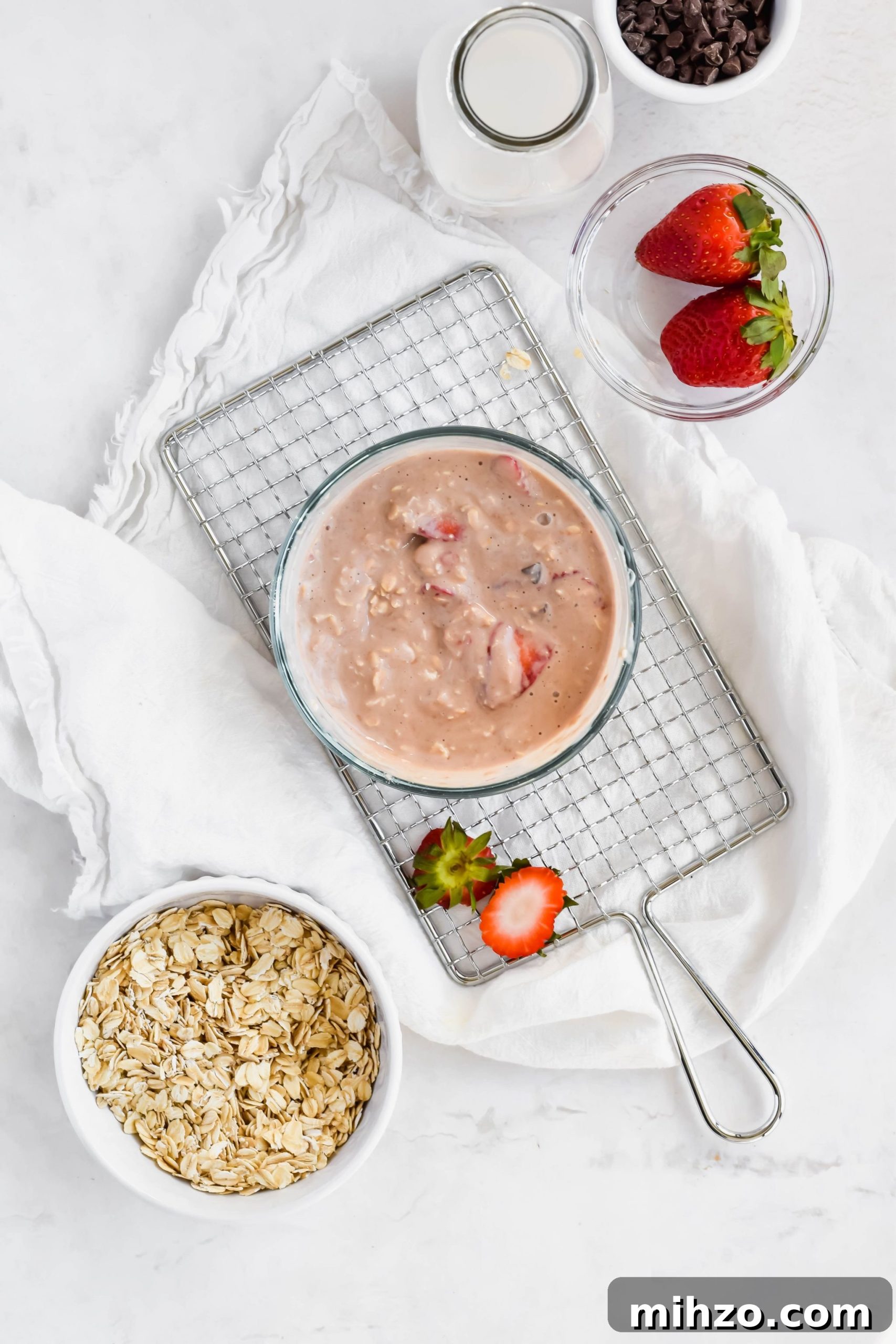 Chocolate-Strawberry Overnight Oats Recipe for Creamy Mornings 7 Strawberry Chocolate Overnight Oats batter in a small glass bowl next to a bowl of rolled oats, a bowl of fresh strawberries, a bowl of mini chocolate chips, and a tall milk jug.