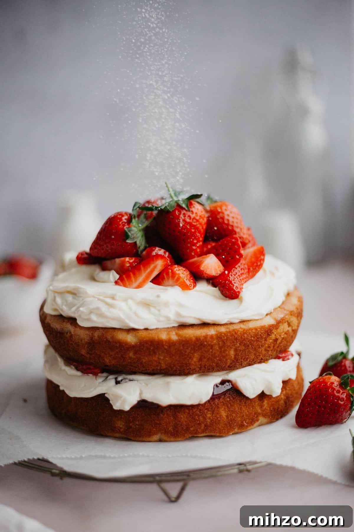 A naked cake, with a whipped cream filling and topping, topped with strawberries. There is a light shower of powdered sugar coming down.
