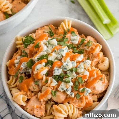A photo of a white ceramic bowl filled with buffalo chicken pasta salad on a marble kitchen counter. The salad is topped with crumbled blue cheese, chopped parsley, and an extra drizzle of buffalo sauce. There's a fork and a napkin beside the bowl. Celery sticks are placed on the side.