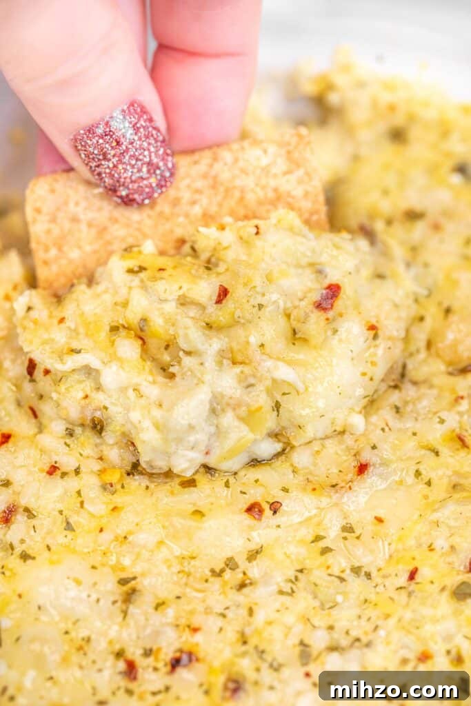 scooping cheesy artichoke dip from a baking dish with a wheat thin cracker