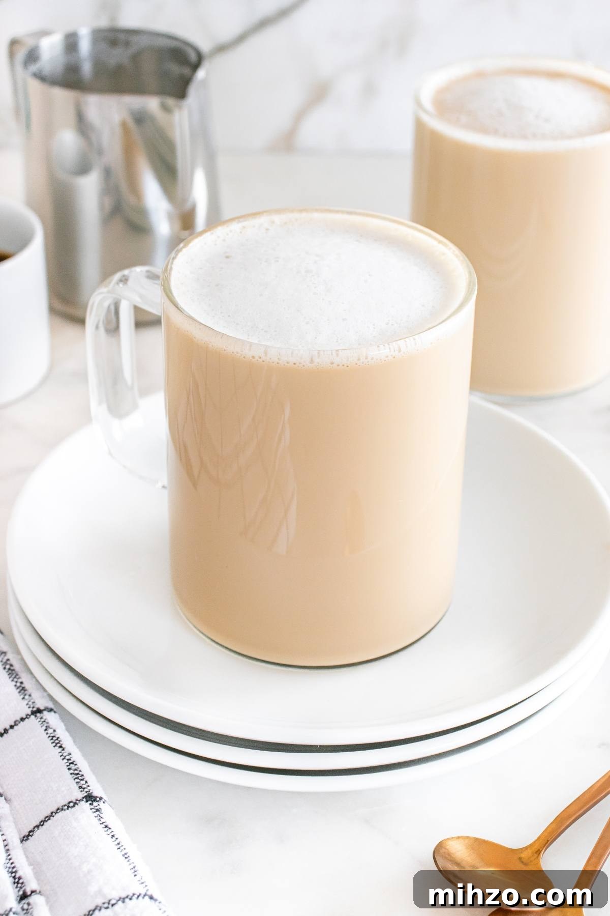 A glass mug of frothy chai latte on a white plate, with another mug and a gold spoon in the background.