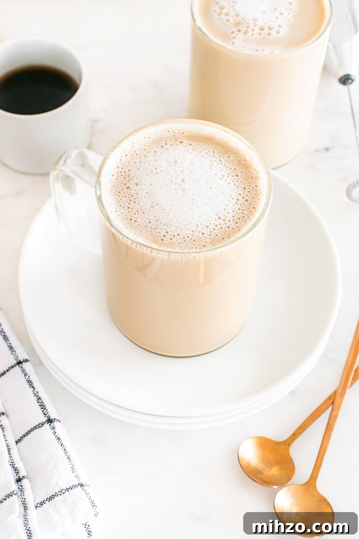 Clear glass mug of frothy coffee on a white plate, with gold spoons and a cup of black coffee nearby.