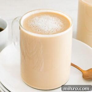 A glass mug of creamy latte breve sits on a white plate with a spoon, next to a cup of black coffee.