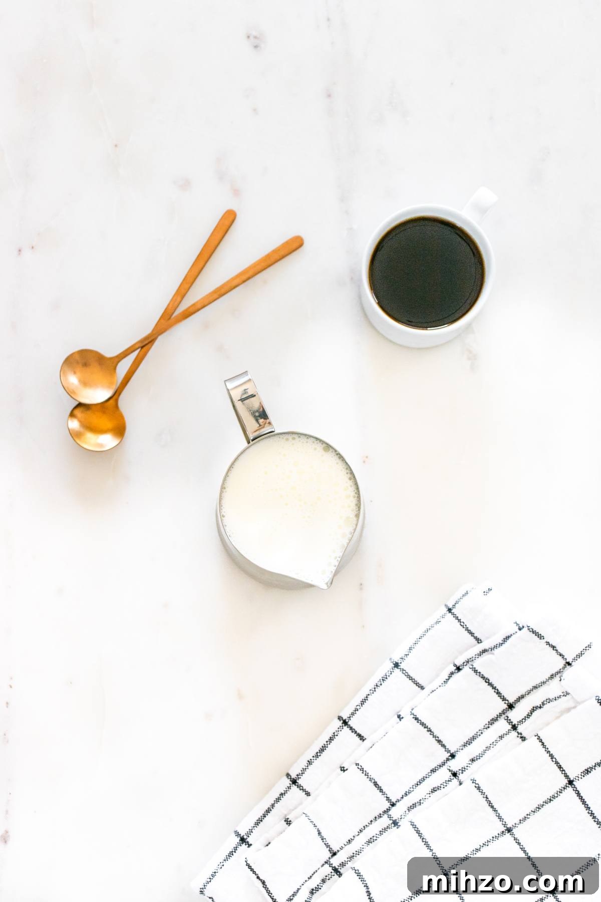 Cup of black coffee, a jug of milk, two golden spoons, and a checkered cloth on a white surface.