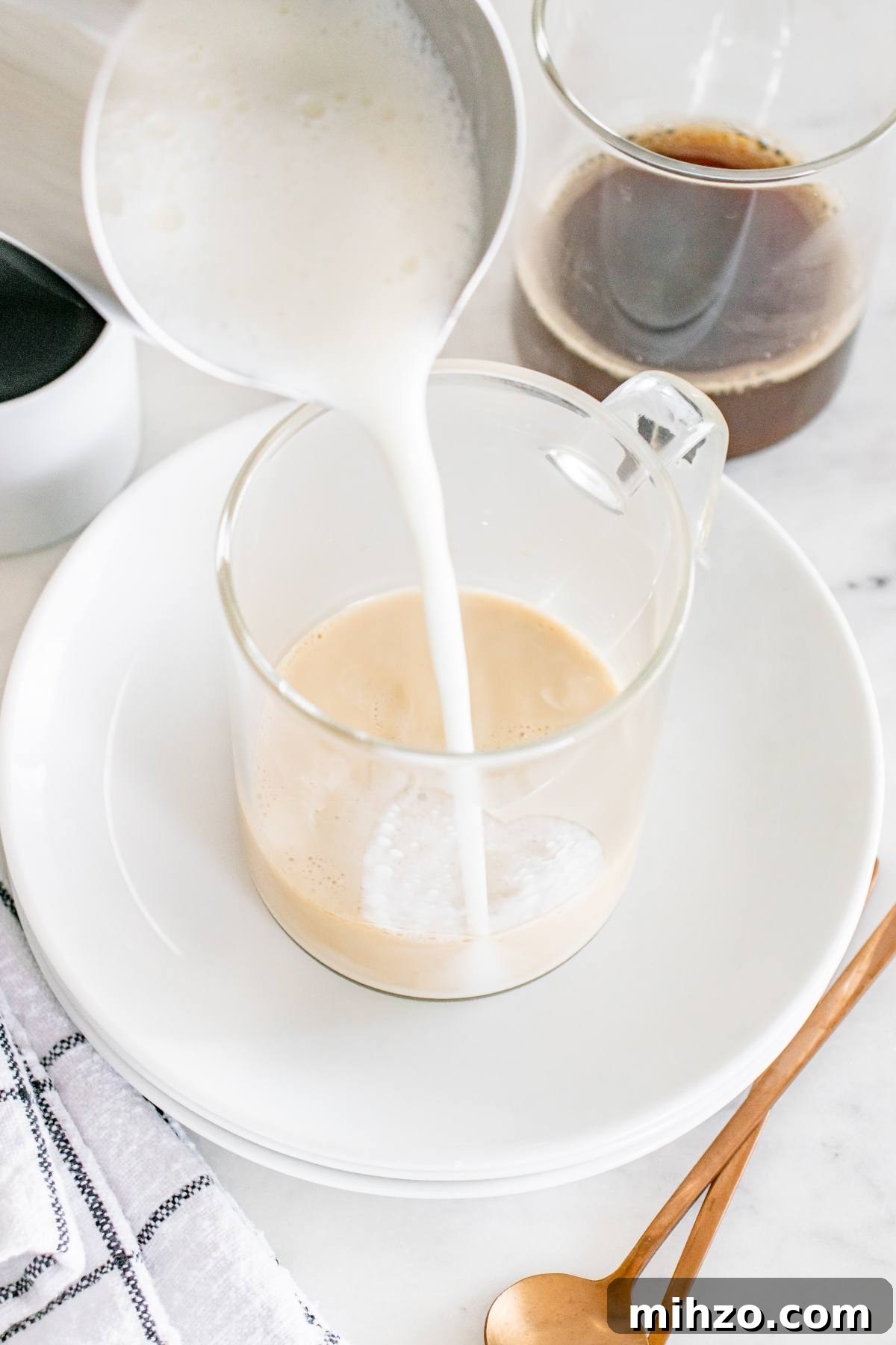 Milk is being poured into a clear glass mug with coffee, on a white plate with a copper spoon nearby.