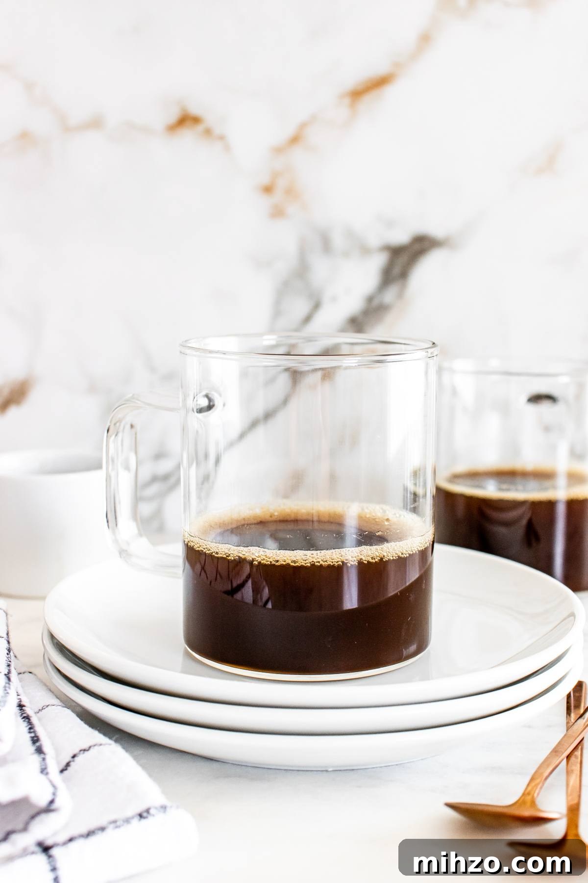 Clear glass mug of black coffee on stacked white plates, with another mug and utensils in the background.