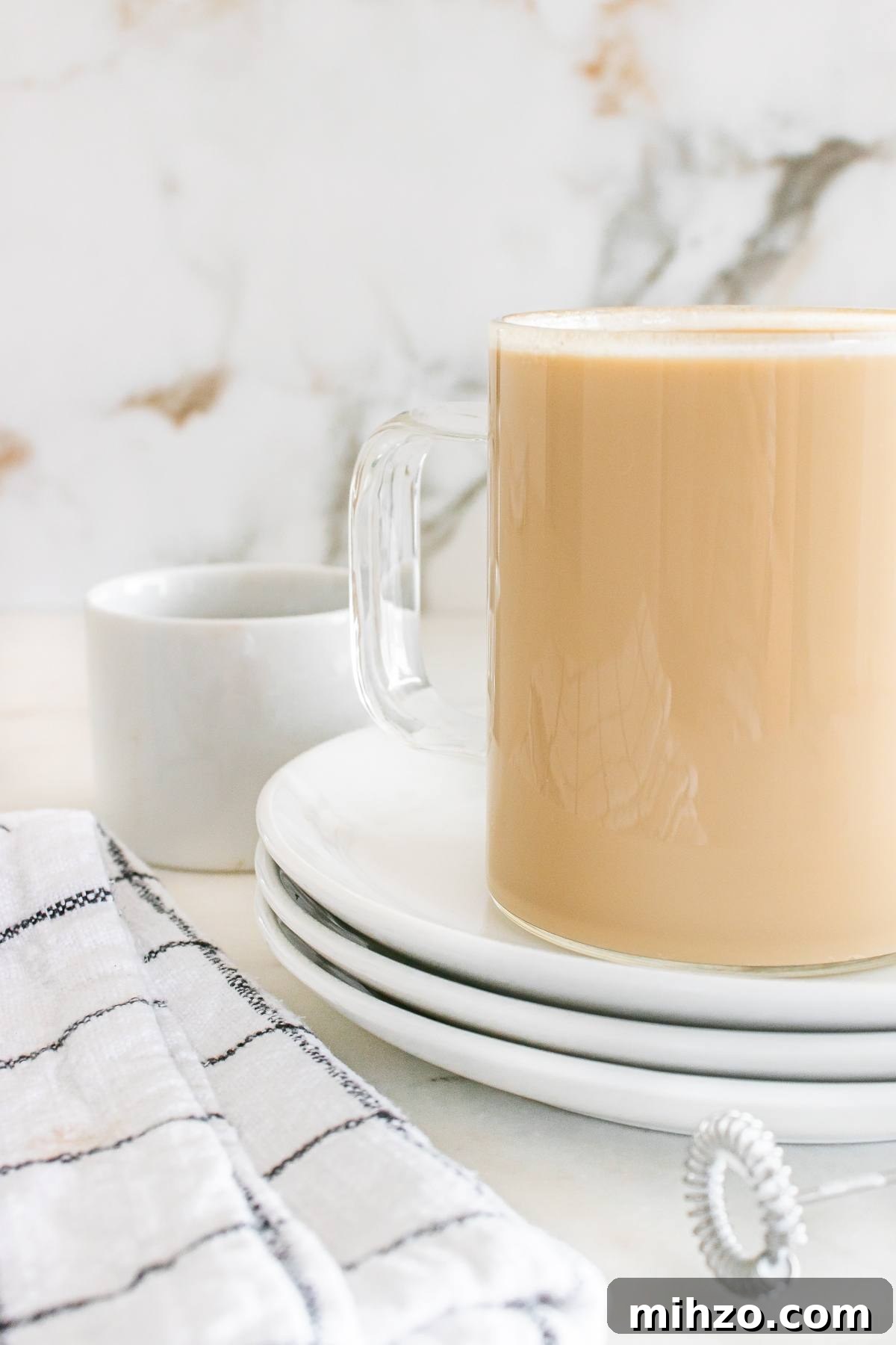A glass mug of coffee with milk on stacked plates beside a small white cup and checkered cloth.