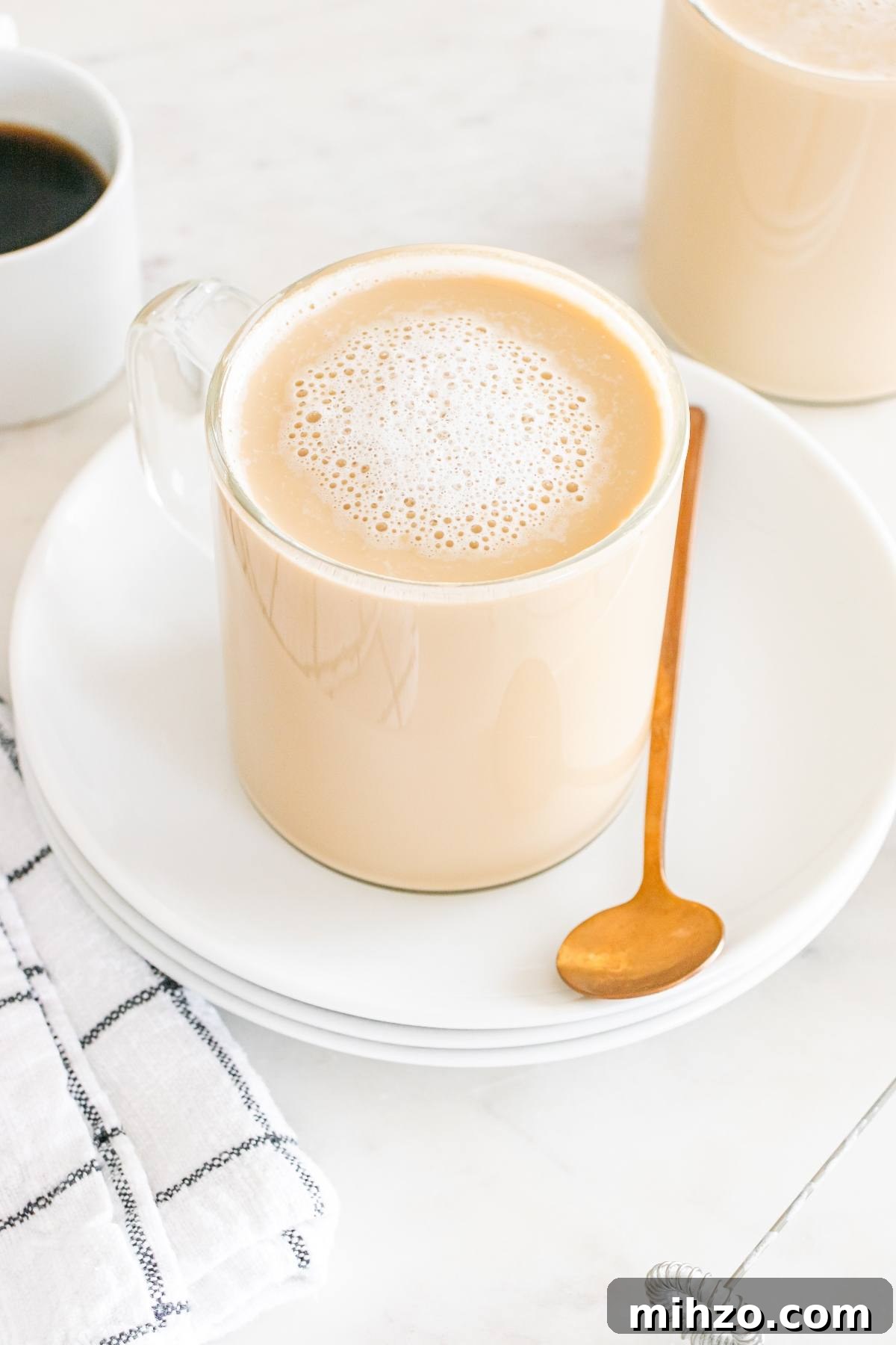 A glass mug of frothy coffee on a white plate with a gold spoon, next to a black-and-white napkin.