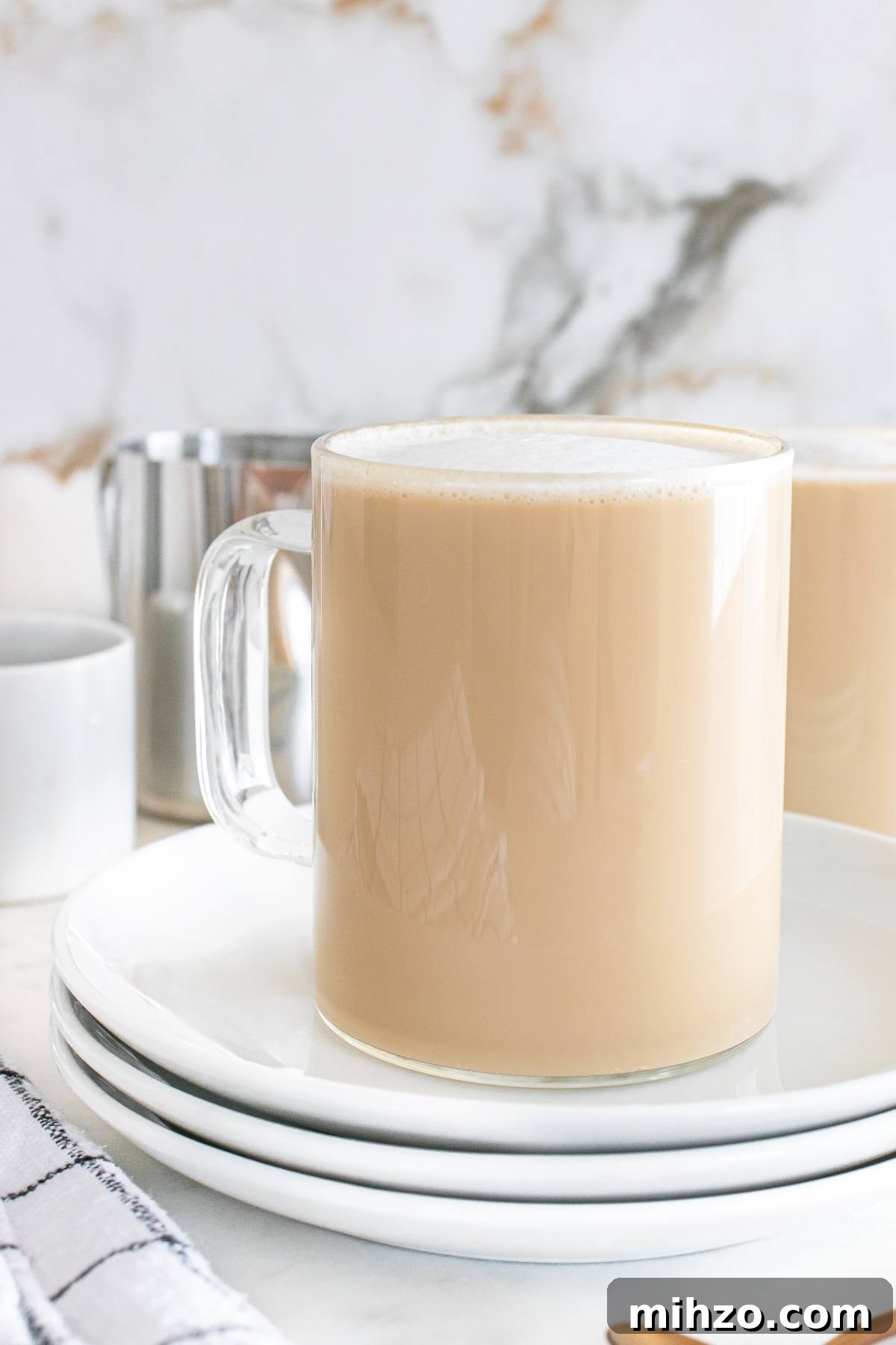 A glass mug of creamy milk tea sits on stacked white plates with a soft, marble background.