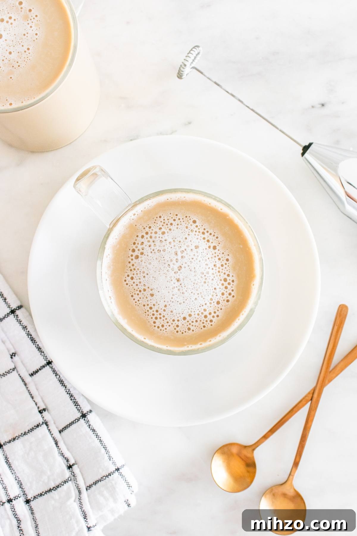 A cup of frothy coffee on a white plate with two gold spoons, a towel, and a milk frother on a marble surface.