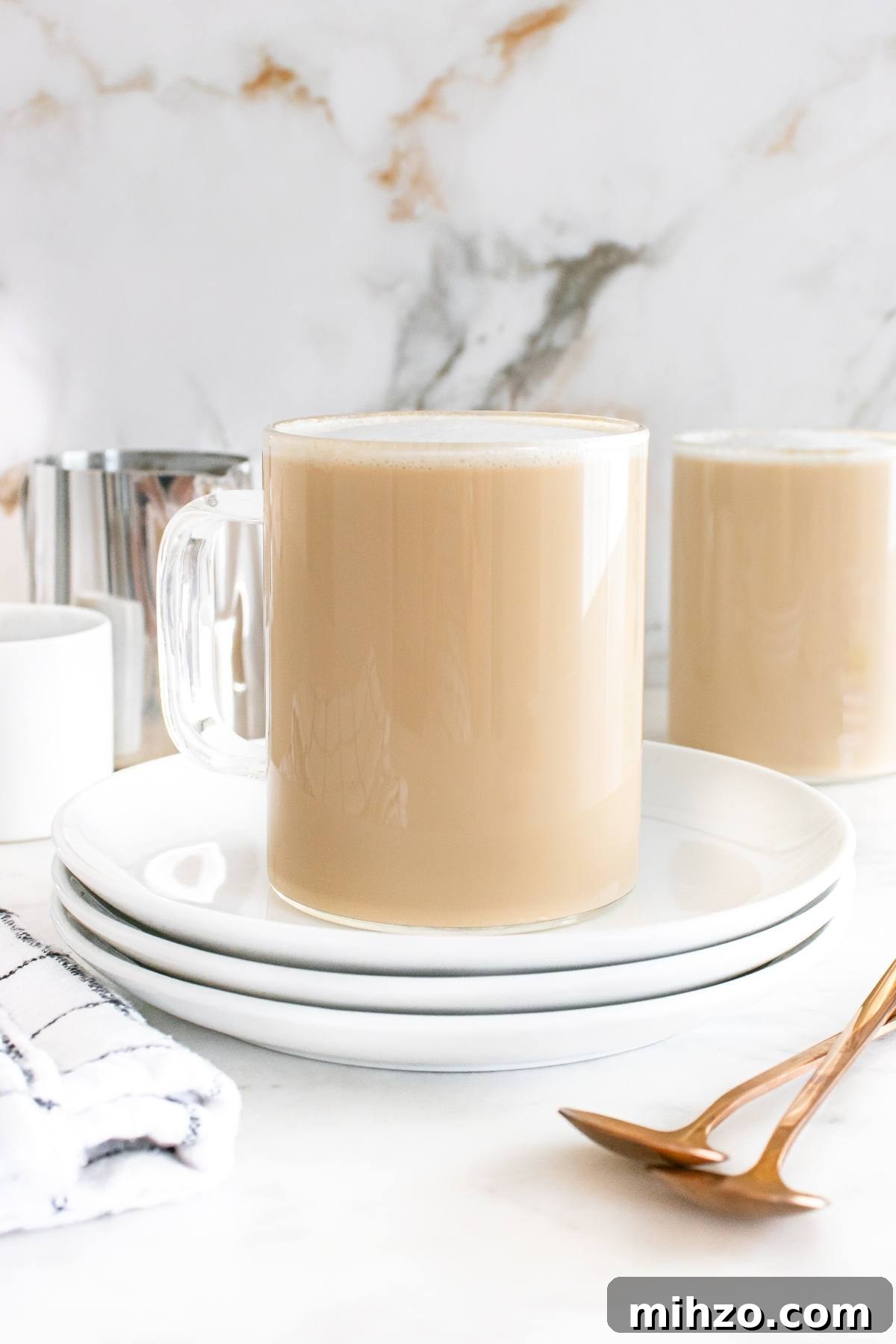 A glass mug of coffee with milk on stacked white plates, next to a napkin and gold spoons.