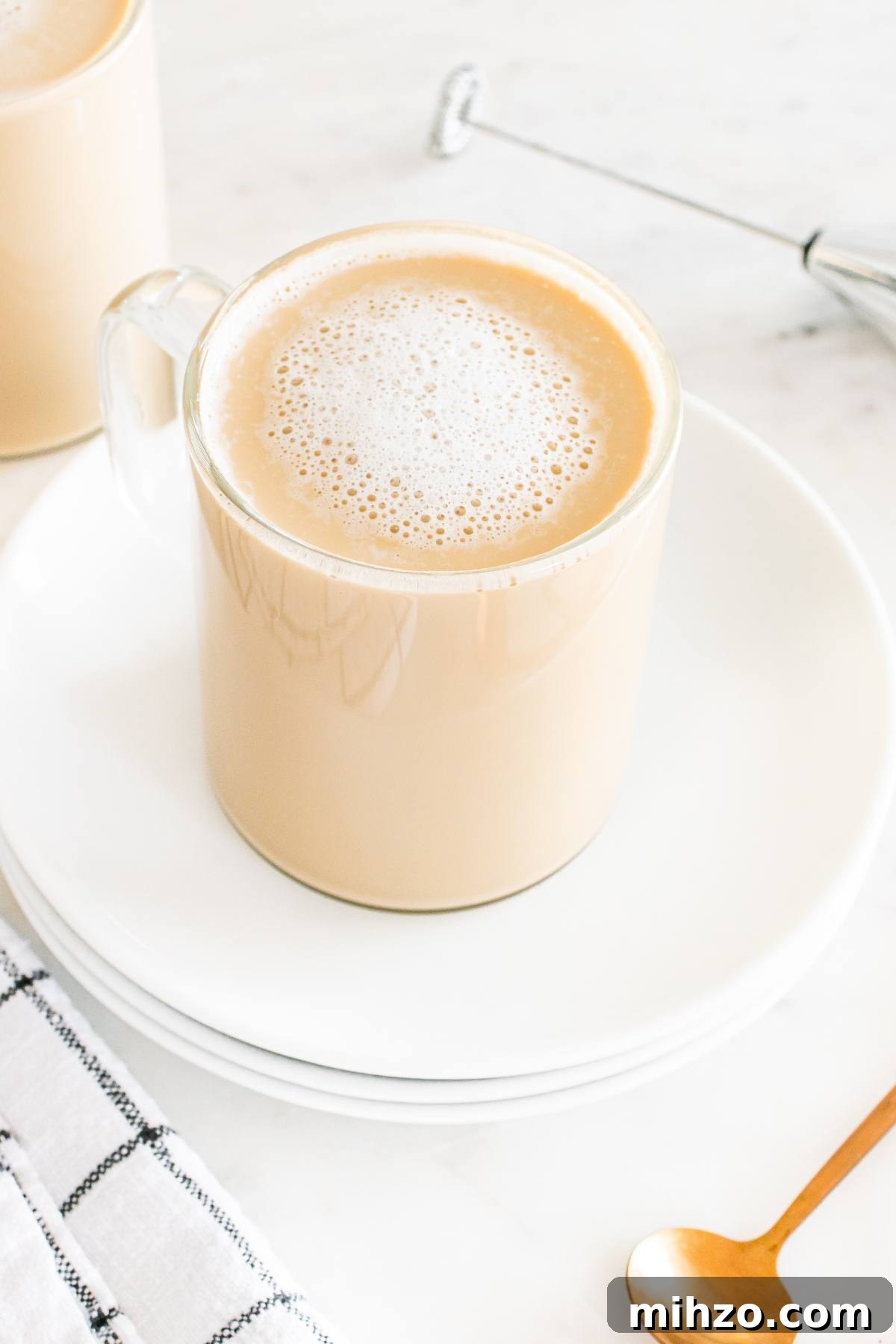 A glass mug of frothy coffee sits on white plates with a gold spoon and a plaid napkin nearby.