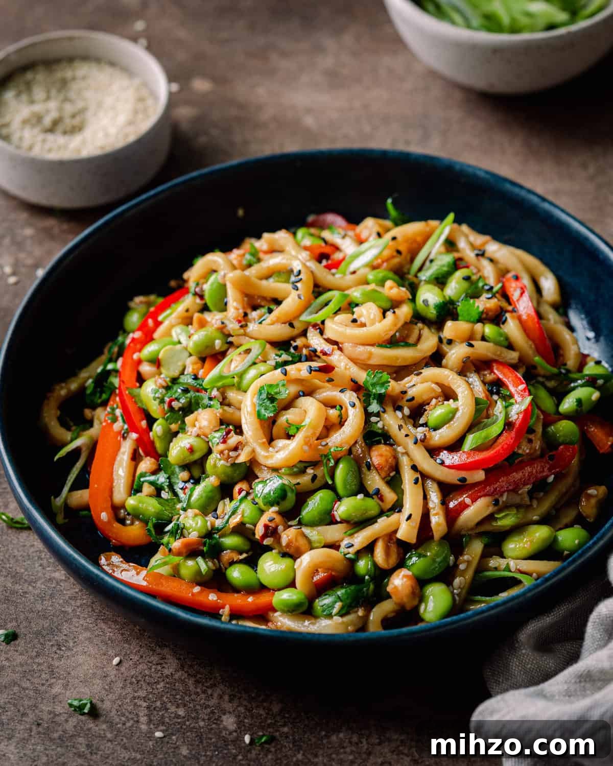 vegan noodles with chili garlic sauce and bell peppers in navy blue bowl on brown backdrop, with bowl of scallions and bowl of sesame seeds