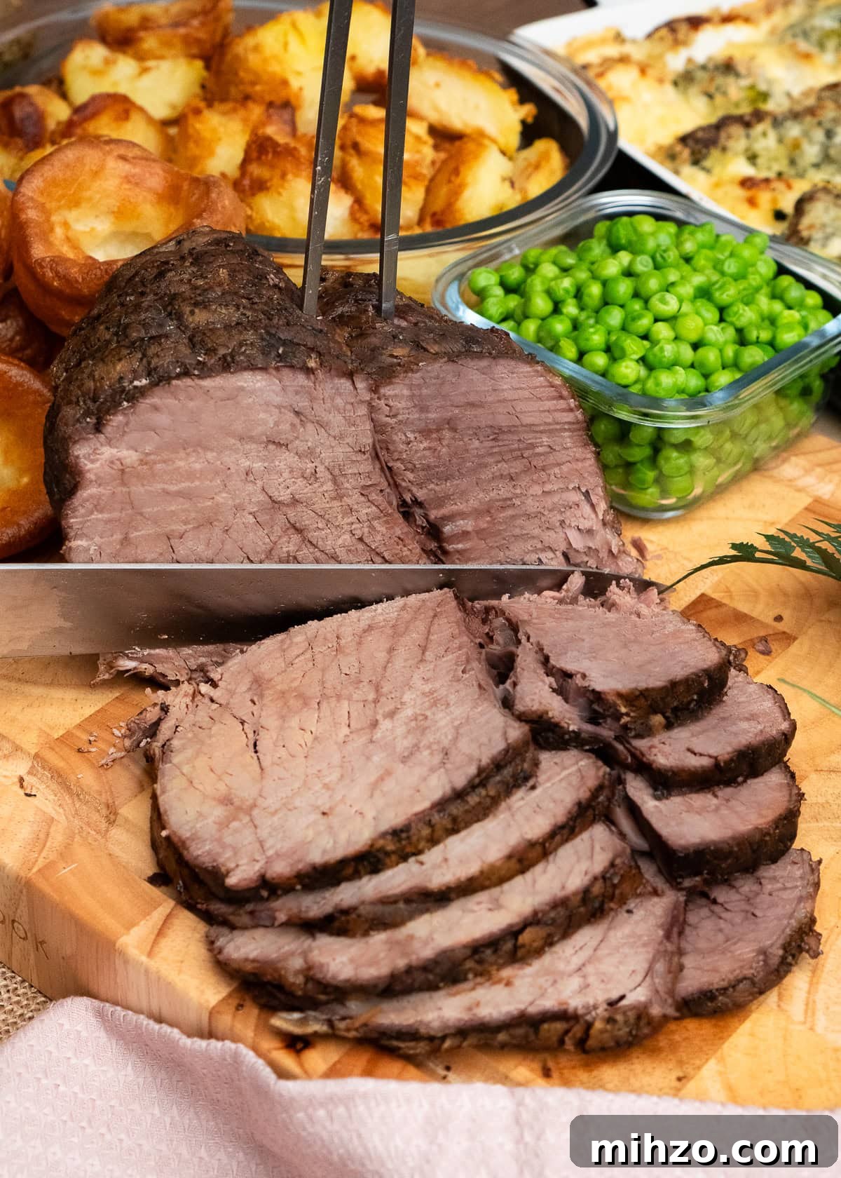 Slicing a slow cooker topside beef joint with a sharp knife and meat fork with peas and roast potatoes in the back ground