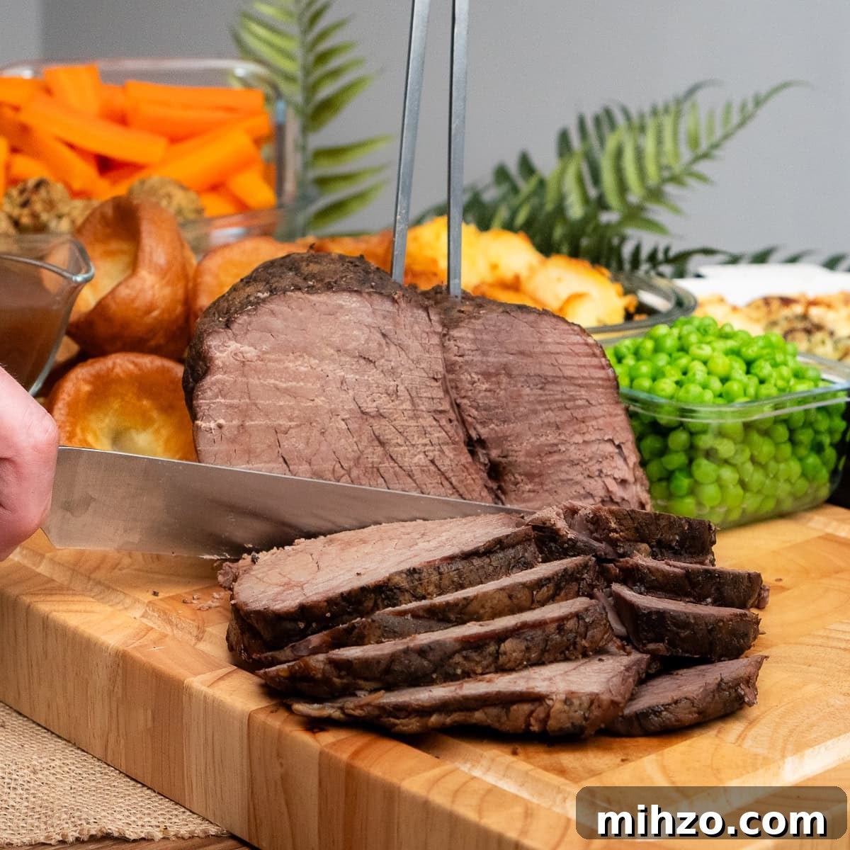 Slicing a slow cooker roast beef joint with a sharp knife and meat fork with peas and roast potatoes in the back ground