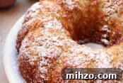 A Jewish Apple Bundt Cake dusted with powdered sugar on a white plate. In the upper left corner is a green and red apple. In the upper right corner is a powdered sugar shaker.