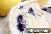 A slice of lemon blueberry bundt cake frosted with lemon cream cheese frosting sitting on a small white plate. Above the plate is a whole lemon and a blueberry lemon bundt cake frosted with lemon cream cheese frosting with a slice taken out of it.