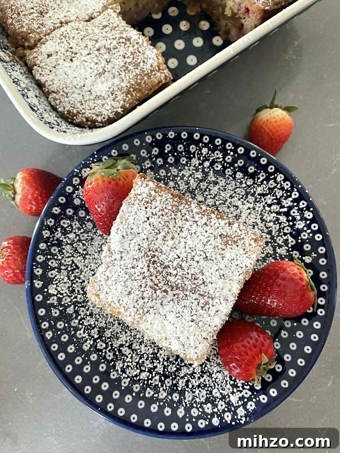 A square piece of strawberry rhubarb cake dusted with powdered sugar on a small dark blue white polka-dotted plate dusted with powdered sugar with two strawberries on the right of the piece of cake and one on the left. A few strawberries are surrounding the top of the plate. The corner of a 9x13 baking dish of a sliced strawberry rhubarb cake is in the upper left middle.