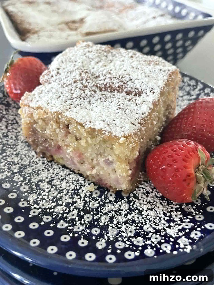 A small stack of dark blue, white polka-dotted small plates with the top plate having dusting of powdered sugar and a slice of strawberry rhubarb coffee cake with two strawberries on the right of the cake and one strawberry on the left of the cake. Faded in the background is a 9x13 baking dish of Strawberry Rhubarb Coffee Cake.