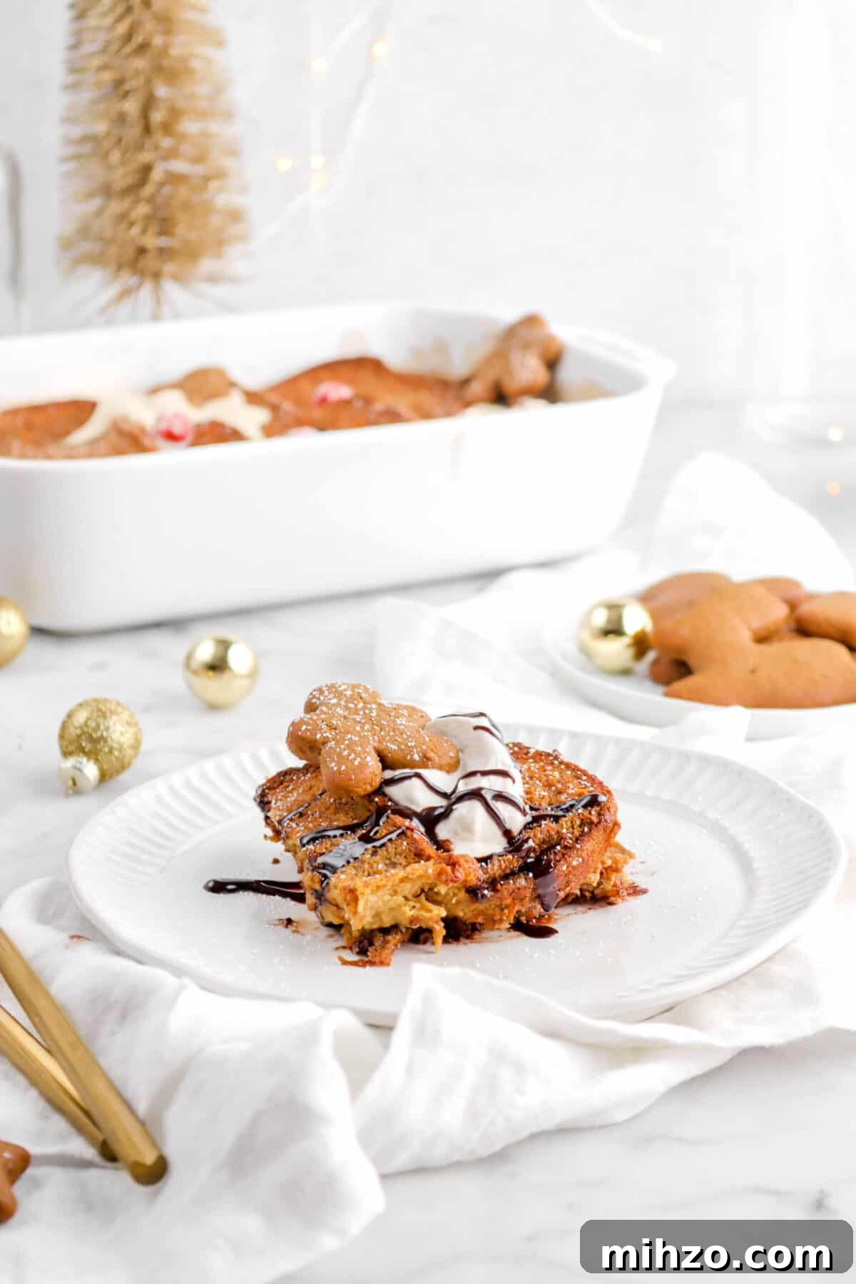 Overnight Gingerbread Chocolate Chip French Toast Bake 7 slice of casserole on white plate with whipped cream, mini cookie, and chocolate drizzle on top with casserole behind
