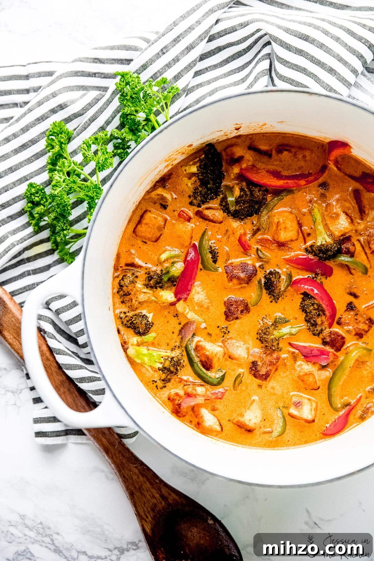 This fragrant Thai red curry is packed with veggies and tender marinated tofu for a satisfying meal. Just add rice or your favorite grain! Overhead view of Thai red curry in enamel Dutch oven with wooden spoon, tea towel, and sprig of parsley