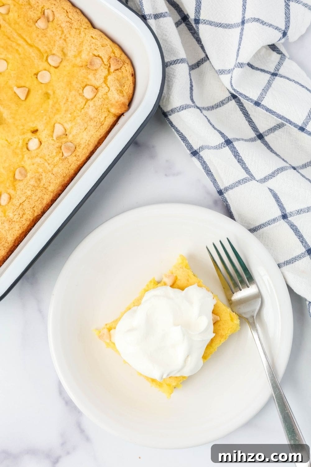 Creamy Lemon Pudding Dump Cake Recipe for Quick Desserts 3 overhead view of a square of low fat lemon cake on a white plate, garnished with reduced-fat Cool Whip. A fork is on the plate, and the remaining pan of cake is next to it.