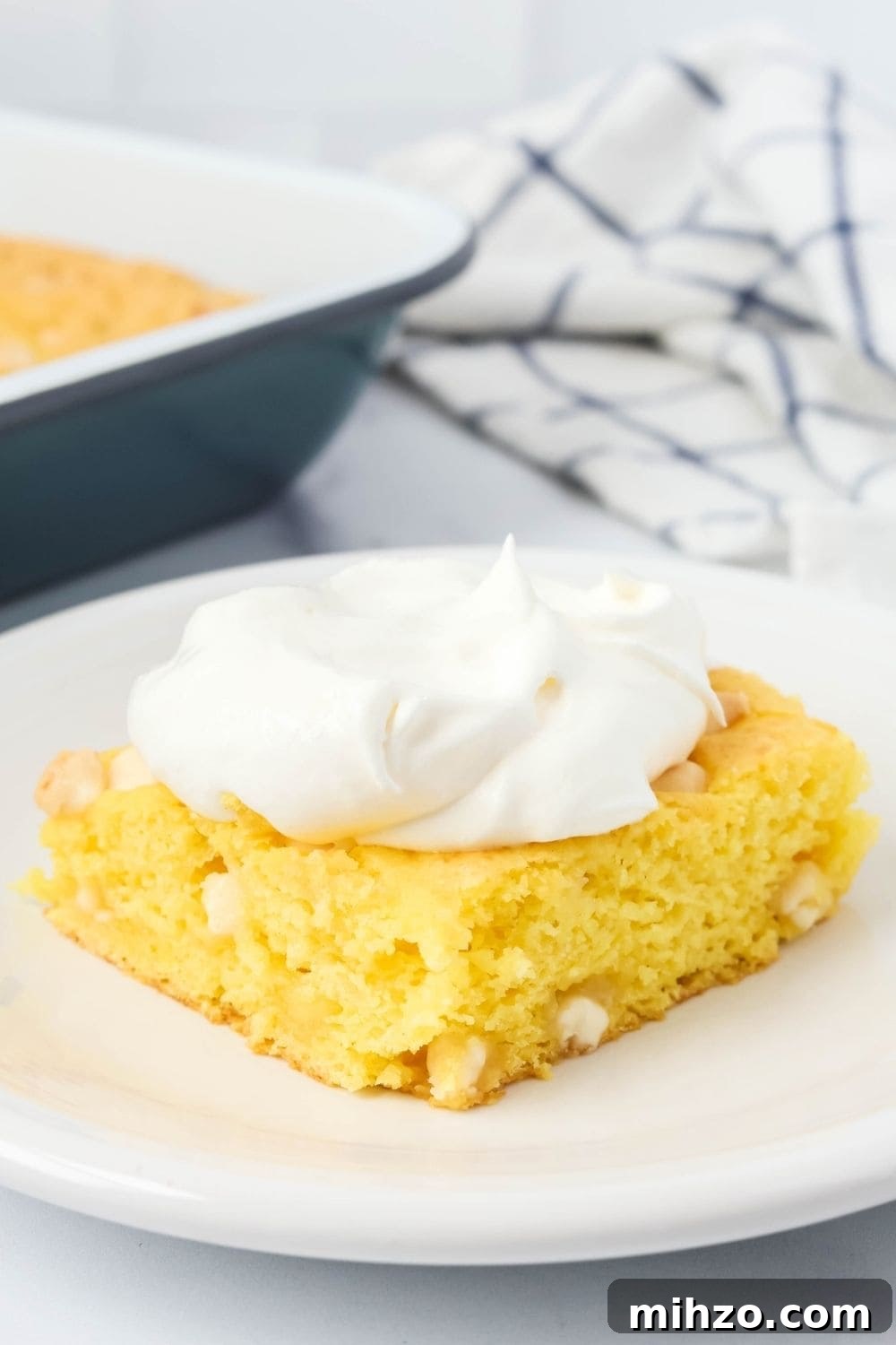Creamy Lemon Pudding Dump Cake Recipe for Quick Desserts 6 close-up view of lemon cake made without eggs or oil. The slice is topped with Cool Whip and served on a white plate. The cake pan is in the background.