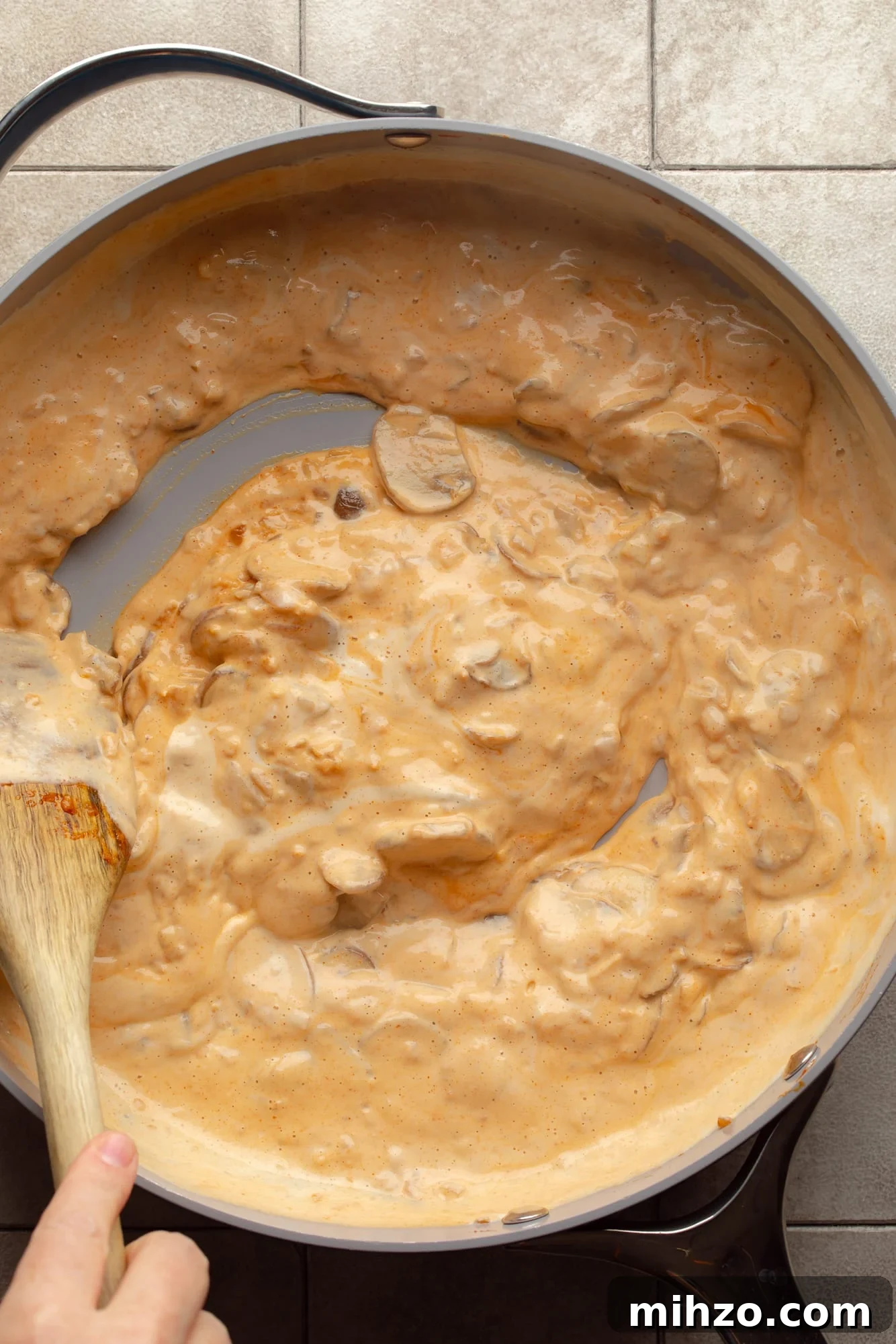 Creamy Gochujang Mushroom Noodles with Sesame and Scallions 5 overhead view of a hand using a wooden spoon to stir a creamy gochujang sauce in a large skillet.
