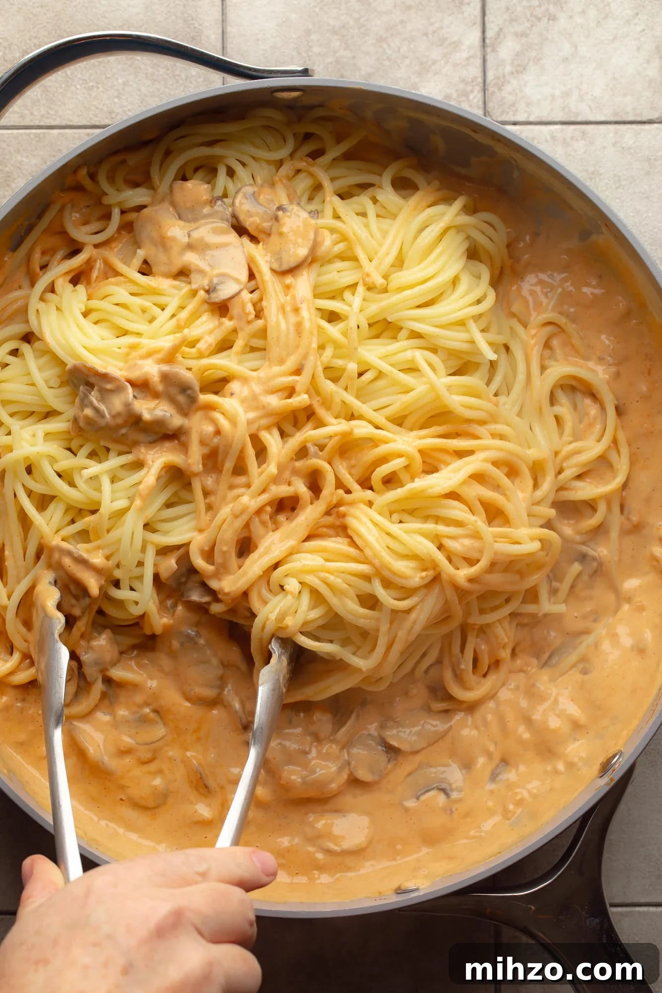 Creamy Gochujang Mushroom Noodles with Sesame and Scallions 6 overhead view of a hand using metal tongs to toss noodles in a creamy gochujang sauce in a skillet.