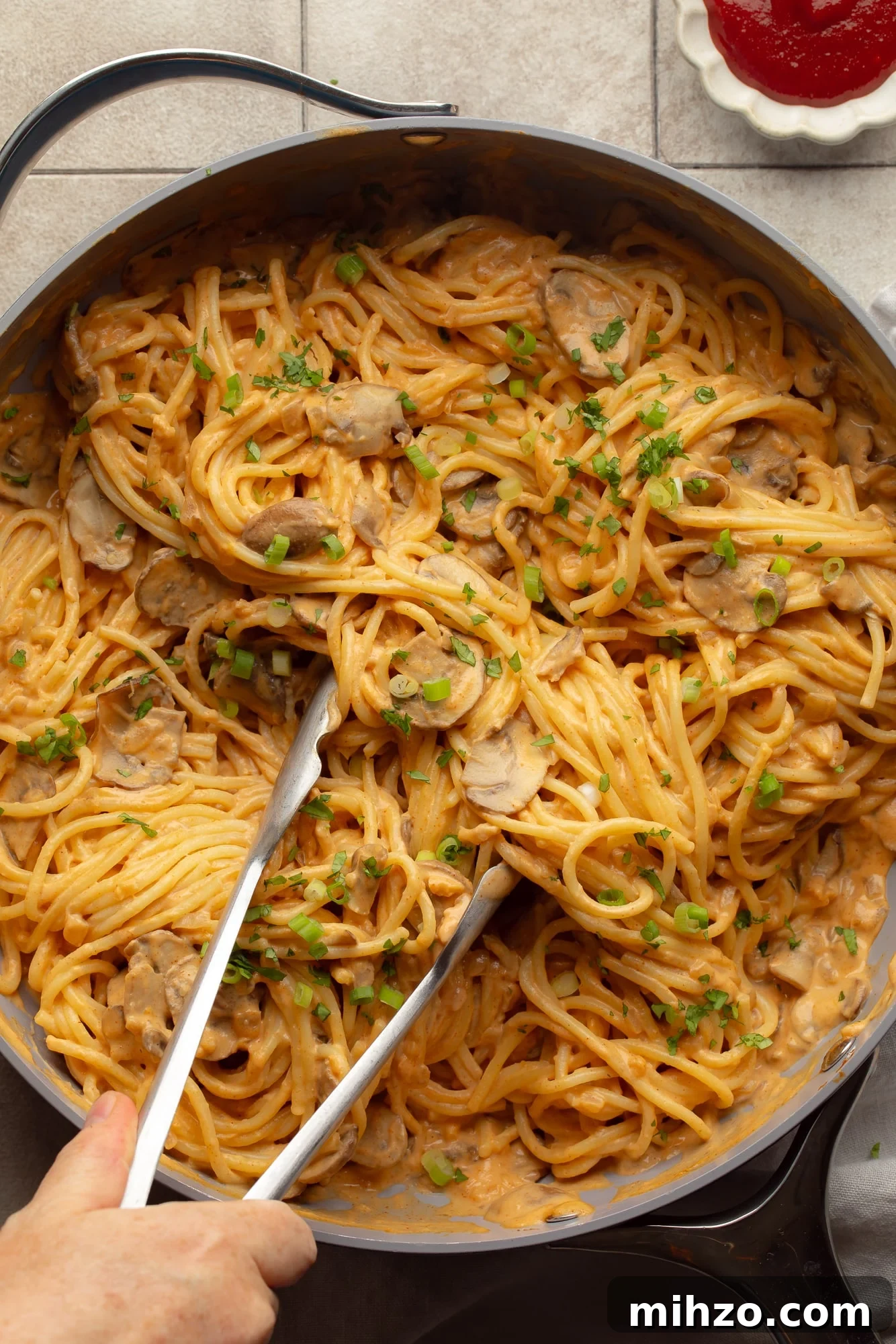 Creamy Gochujang Mushroom Noodles with Sesame and Scallions 7 overhead view of a hand holding metal tongs in a skillet full of creamy gochujang noodles.
