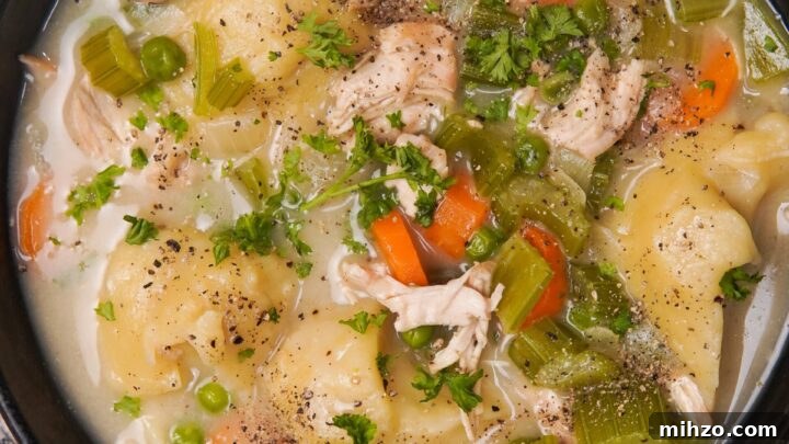 A black bowl filled with gluten-free dumplings, shredded chicken, carrots, celery, peas, and broth, topped with chopped parsley and black pepper.