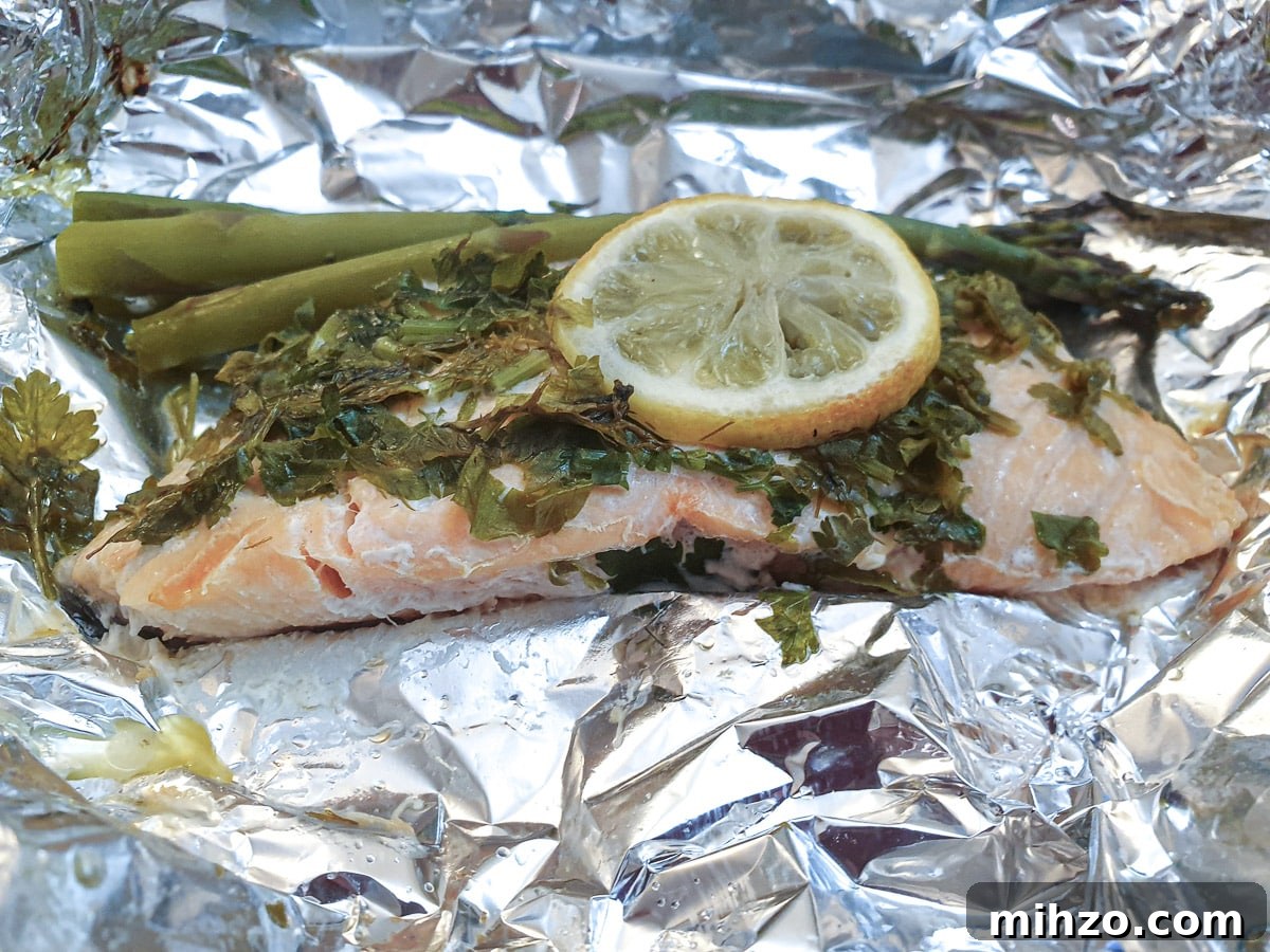 Lemon Herb Butter Salmon and Asparagus Parcels 3 Close up of a piece of cooked salmon on a sheet of tin foil, covered with herbs and lemon, with asparagus spears.