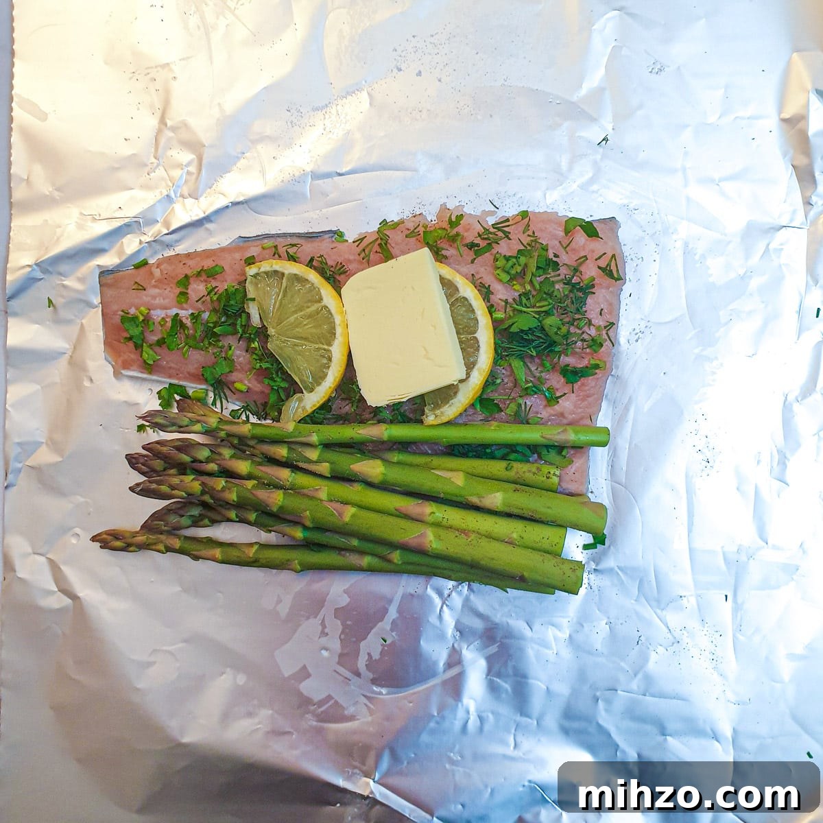 Lemon Herb Butter Salmon and Asparagus Parcels 5 A piece of uncooked salmon topped with herbs, lemon and a knob of butter on a sheet of tin foil.