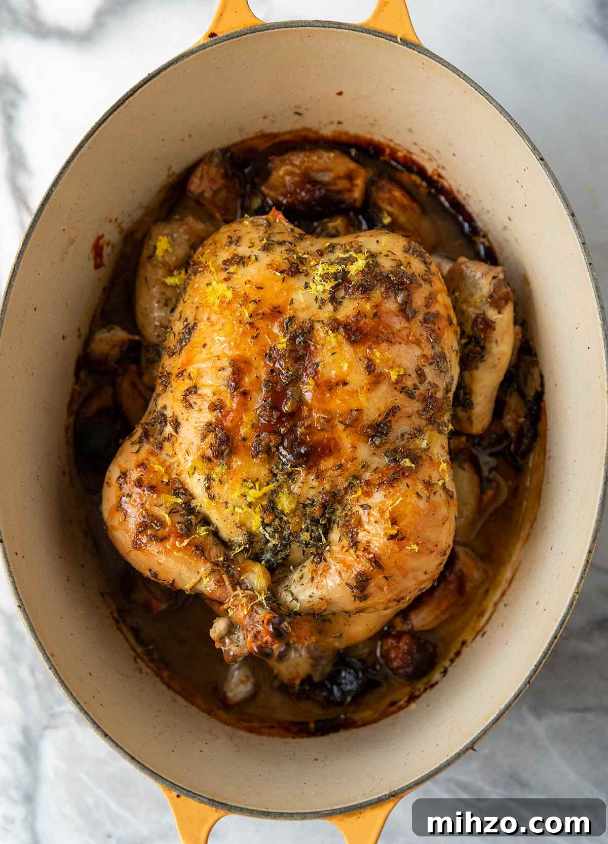 8 Flavorful Whole Roast Chicken Recipes for Every Occasion 5 A large yellow Dutch oven with a roast chicken on a bed of cooked shallots, topped with lemon zest and herbs.