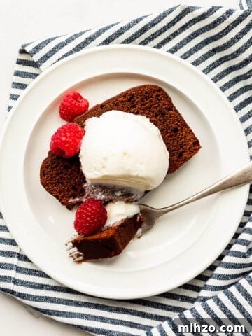 Slice of chocolate loaf cake with a scoop of ice cream and fresh raspberries on a plate with a fork, with a striped linen tucked underneath.
