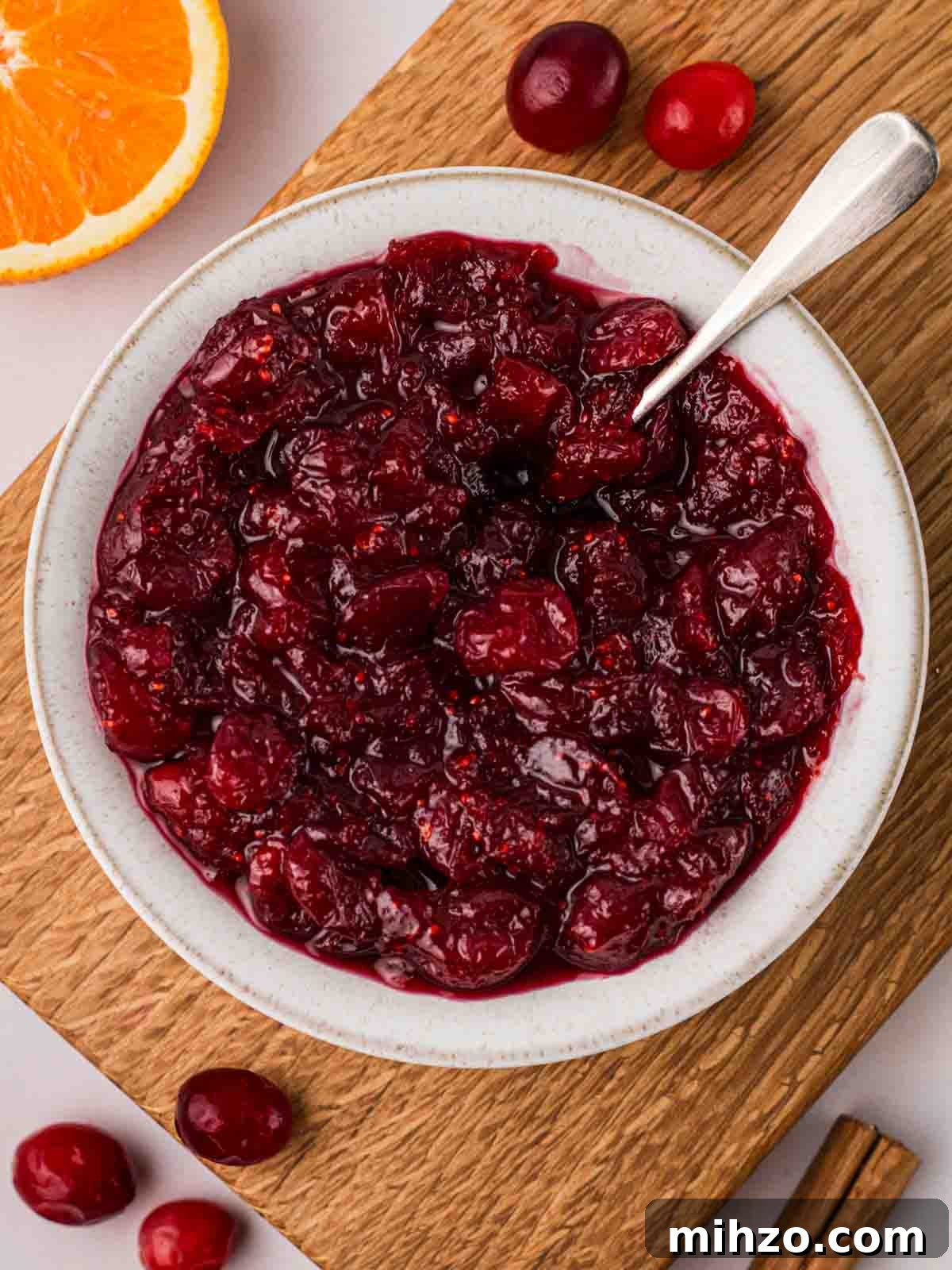 A board with a white bowl filled with cranberry sauce, with cranberries and an orange at the side.