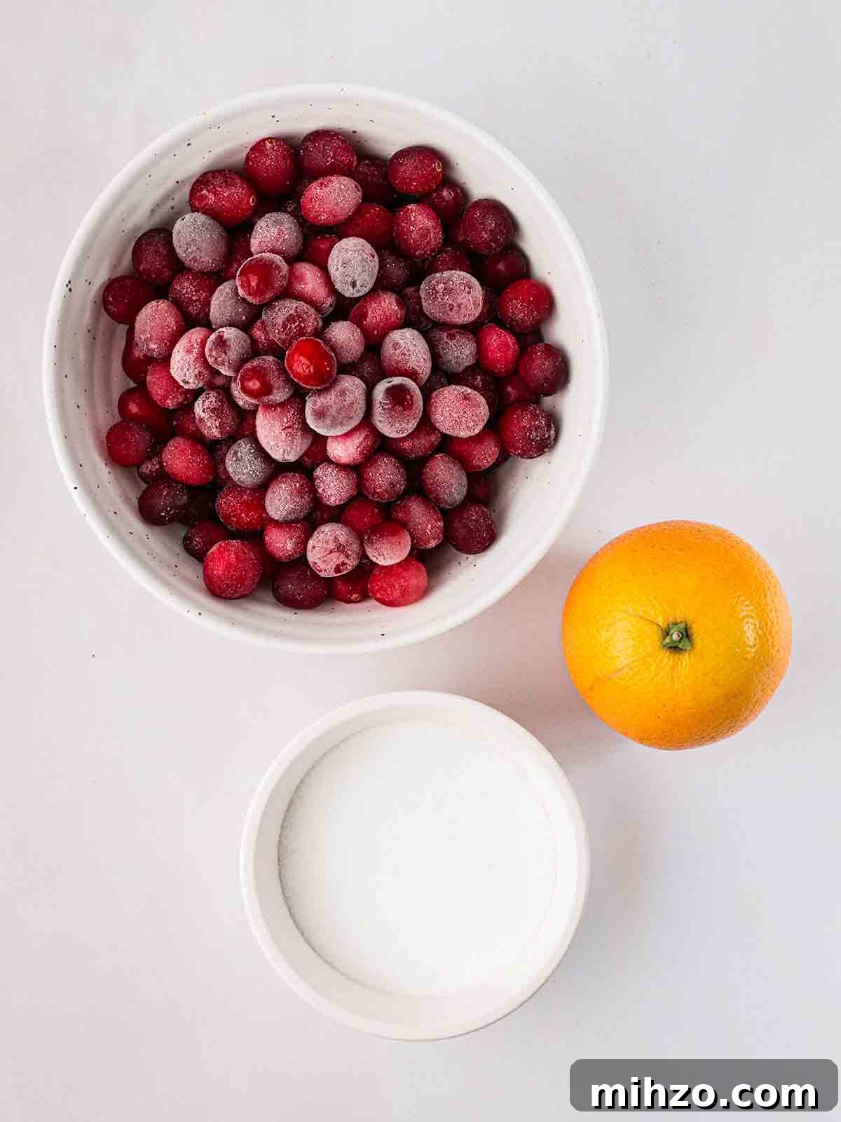 The ingredients for making cranberry sauce laid out on a counter top, including a bowl of frozen cranberries, an orange and sugar.