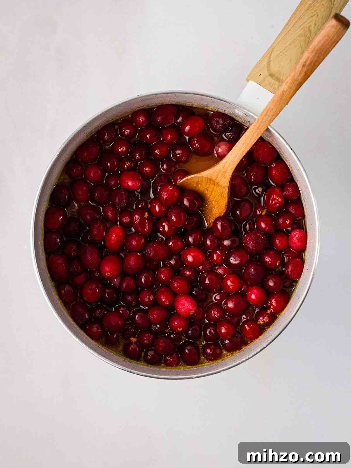 A pan with a wooden spoon stirring cranberries for step 1 in the recipe for Cranberry Sauce.