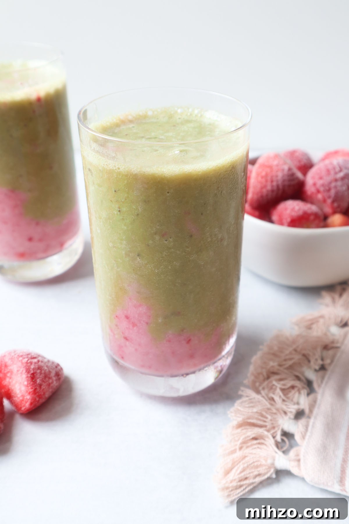 two glasses with strawberry matcha smoothie with a pink strawberry layer at the bottom and a green matcha layer on top with a bowl of strawberries behind the smoothie.