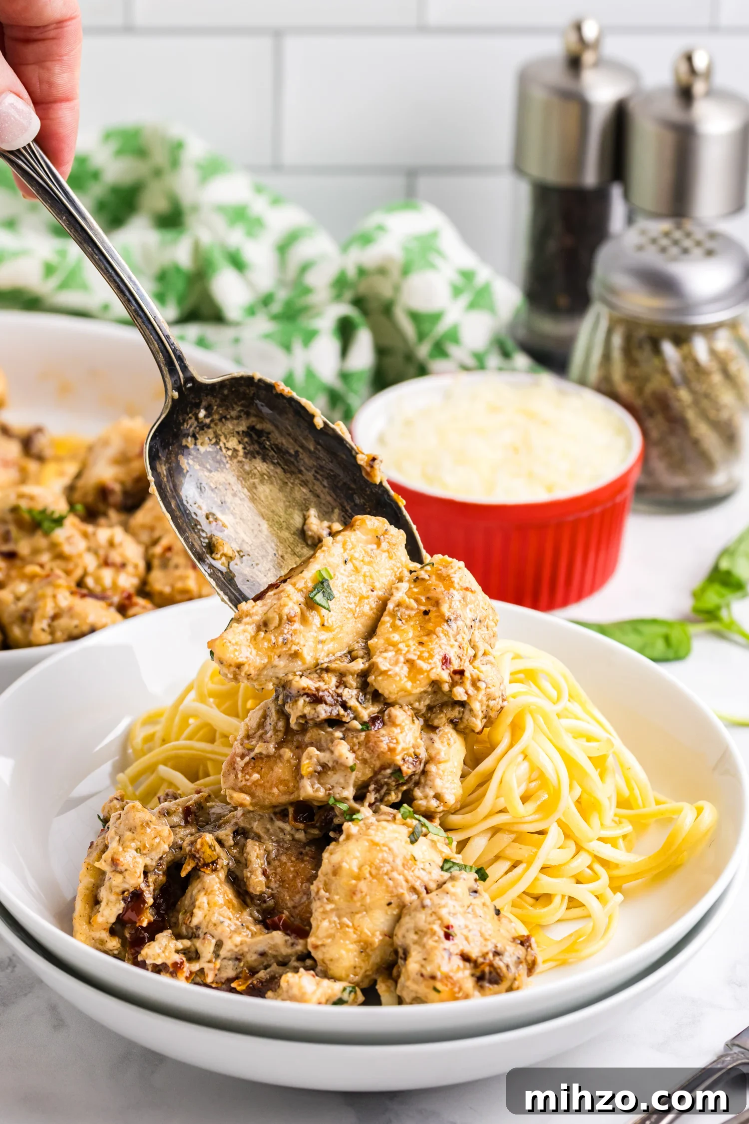 A spoon lifts creamy sun-dried tomato chicken over a bowl of cooked pasta, with grated cheese and seasonings visible in the background.