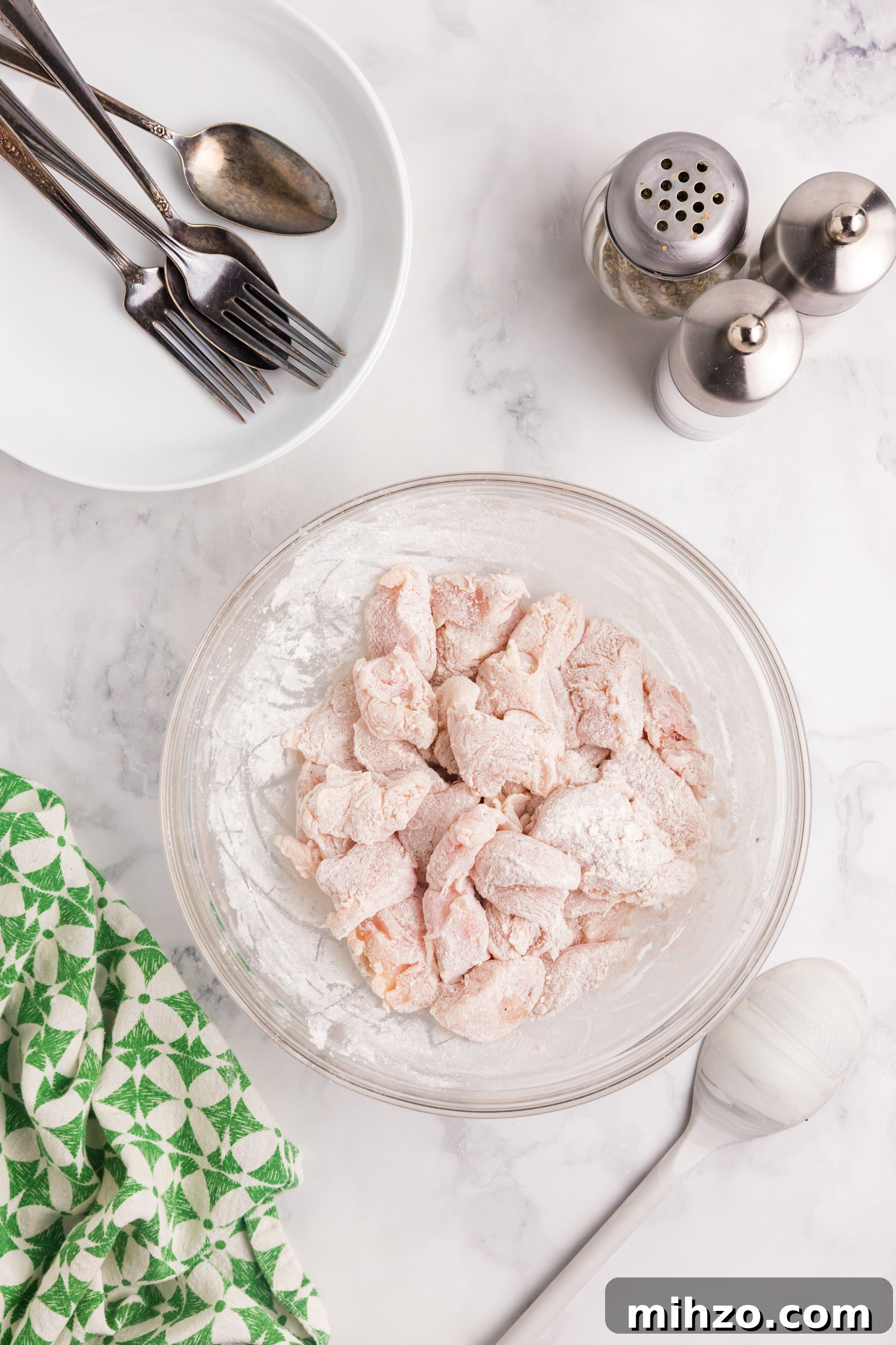 Raw chicken pieces coated in flour sit in a glass bowl on a marble countertop