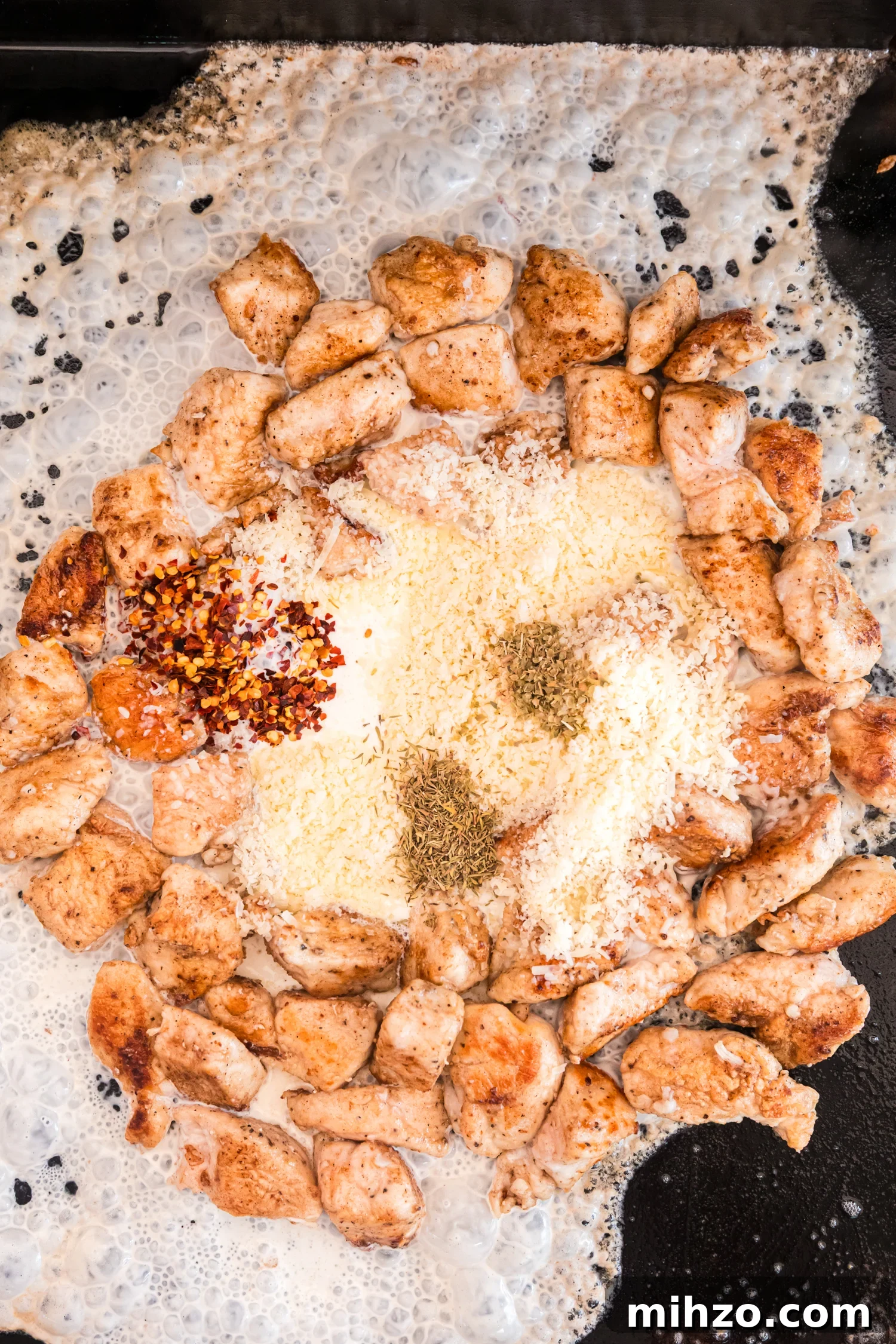 Cooked chicken pieces are arranged in a circle on a griddle while cream, grated cheese, red pepper flakes, and dried herbs melt together in the center to form a sauce.